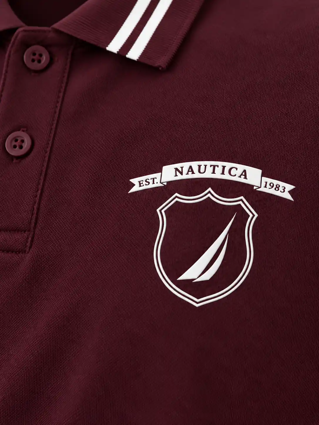 Detail of white raised high-density chest printing with crest outlines, banner lettering and a sail-like graphic on burgundy fabric